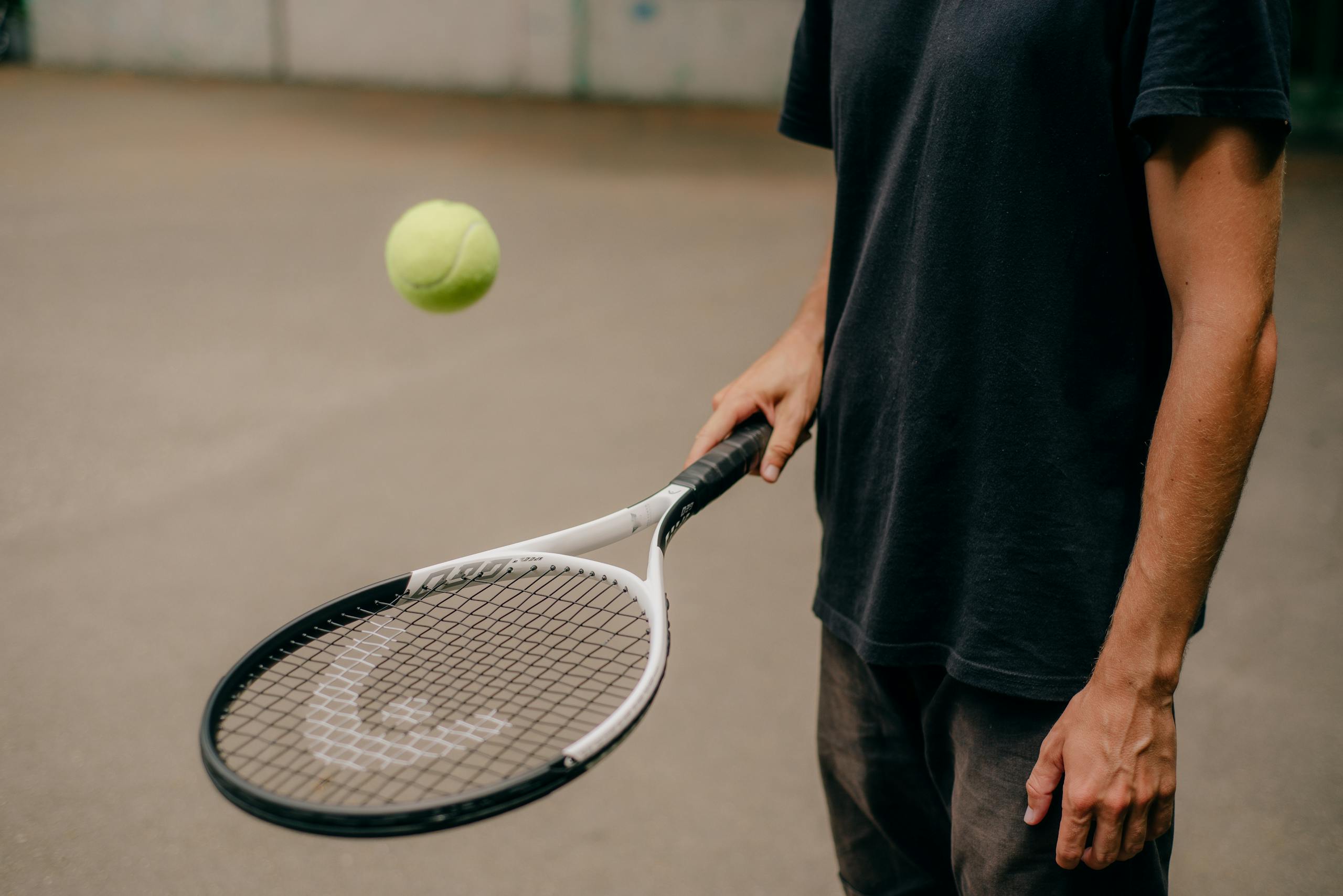 A man in a black t-shirt skillfully handling a tennis racket with a ball in motion outdoors.